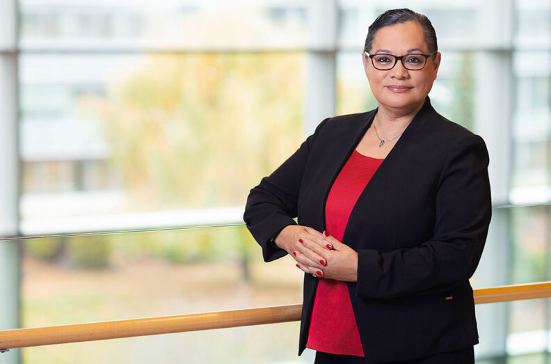 Gloria Ochoa-Bruck in a black blazer and red top, standing indoors by a wooden railing.