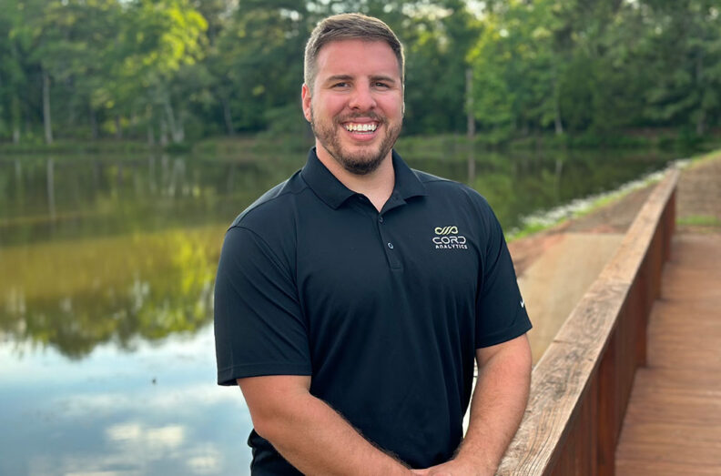 John Howerton in a black CORD Analytics polo shirt standing on a wooden path near water and trees.