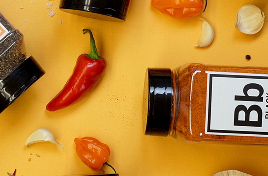 Assorted spices and peppers on a yellow background, including jars, chili, garlic, and peppercorns.