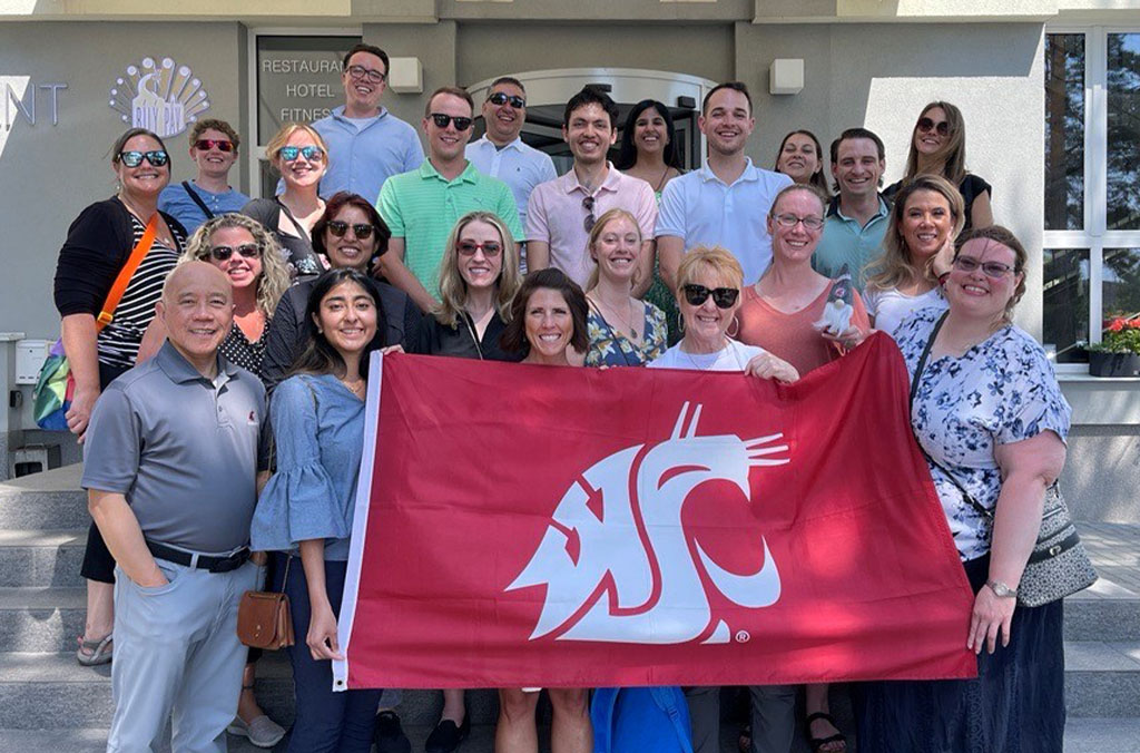 MBA group in Prague holding a WSU flag on steps in front of a building with restaurant and hotel signs.
