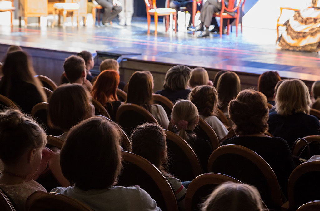 Audience seated in a theater watching a stage performance with chairs and performers.