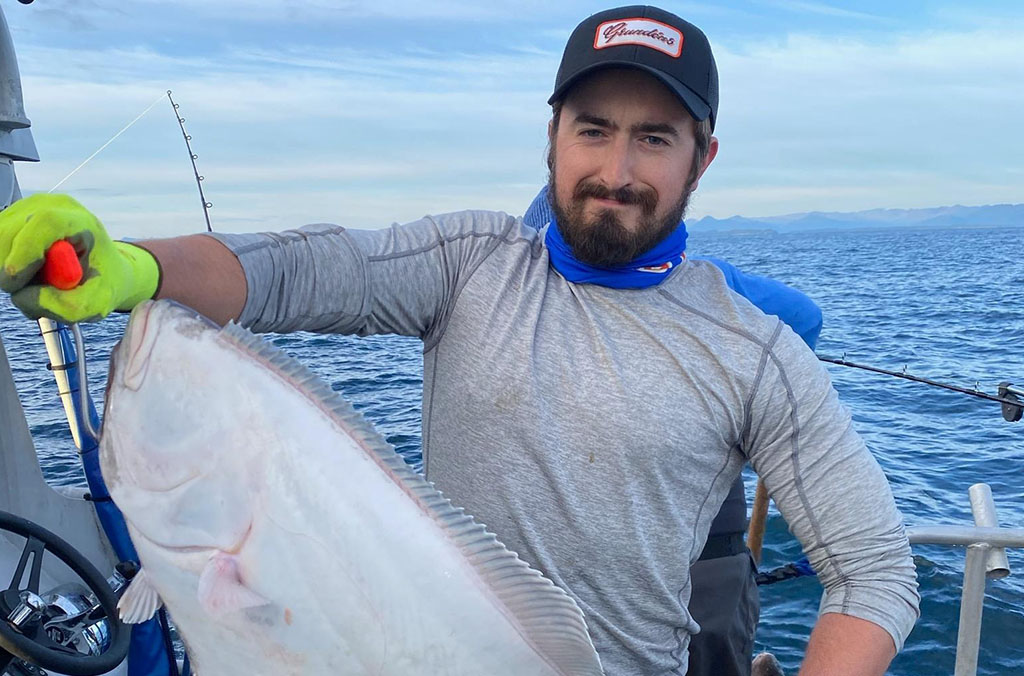 Sean Russell on a boat holding a large fish, wearing green gloves and a black cap, with the ocean in the background.