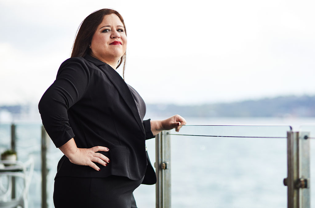 Suellen Dos Santos Frank in a black outfit standing on a balcony overlooking water and distant hills.