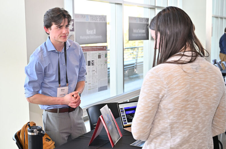 Aidan Finnegan (left) speaks with an attendee at the BPC Trade Show, with a laptop and informational posters in the background.
