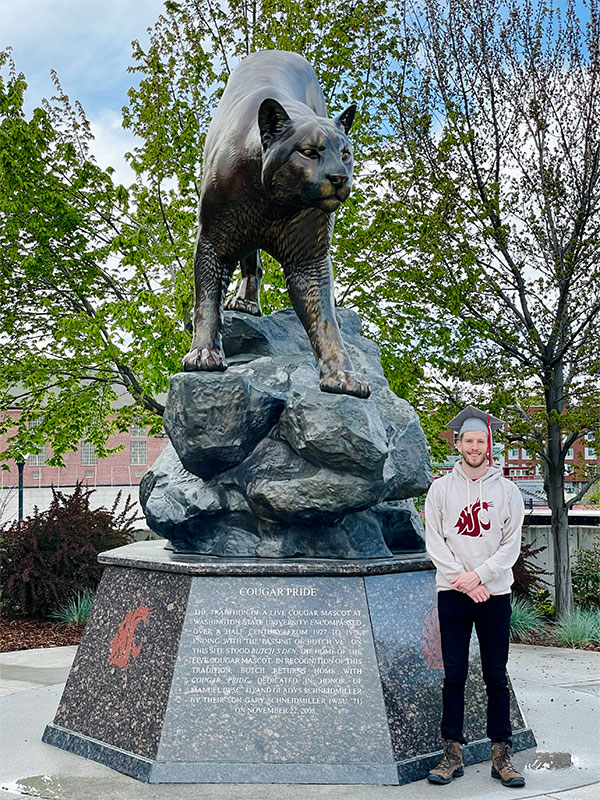 OMBA graduate Jeff Browne wearing a graduation cap and Cougar head logo sweater, standing in front of the 'Cougar Pride' statue on the Pullman campus, with trees and buildings in the background.