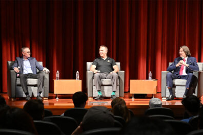 Three panelists seated on stage in gray armchairs during the Business Technology Symposium, with a red curtain backdrop and small tables holding water bottles between them. Audience members are visible in the foreground.