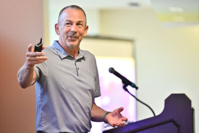 Joe Valacich presenting at the Hubman Lecture, standing beside a podium with a microphone and gesturing while holding a presentation remote. A blurred projection screen is visible in the background.