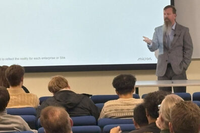 Collin Sower speaking during the Walton Lecture, standing in front of a large projection screen. Audience members are seated in rows of blue chairs in the foreground.