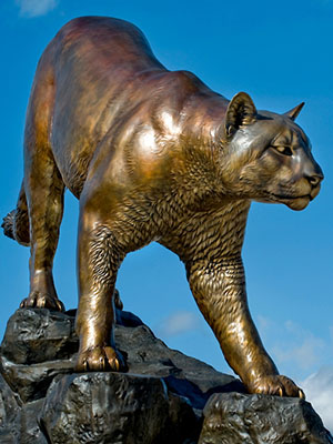 Bronze “Cougar Pride” statue of a cougar standing on rugged rocks against a blue sky.