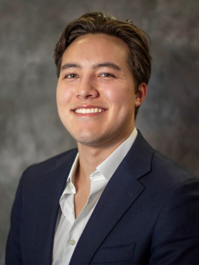 Grant Shinohara wearing a dark navy blazer over a white collared shirt, posed against a mottled gray studio backdrop.