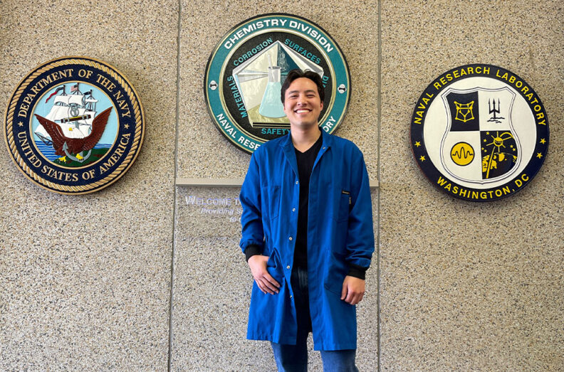 Grant Shinohara wearing a blue lab coat, standing in front of three circular emblems for the Department of the Navy, Naval Research Laboratory, and Chemistry Division on a speckled wall.
