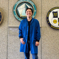 Person wearing a blue lab coat standing in front of wall plaques for the Naval Research Laboratory and Chemistry Division, indicating a research or internship setting.
