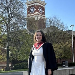 Kaila Chang Freitas wearing a graduation gown and red stole, standing outdoors near the WSU clock tower on a sunny day.
