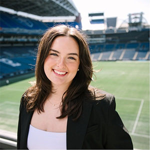 Molly Olney wearing a black blazer over a white top, standing on an outdoor balcony overlooking a large sports stadium with green turf and blue seating sections.