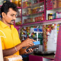 Person in a bright yellow shirt using a card reader at a small shop counter, with shelves of colorful packaged goods in the background.