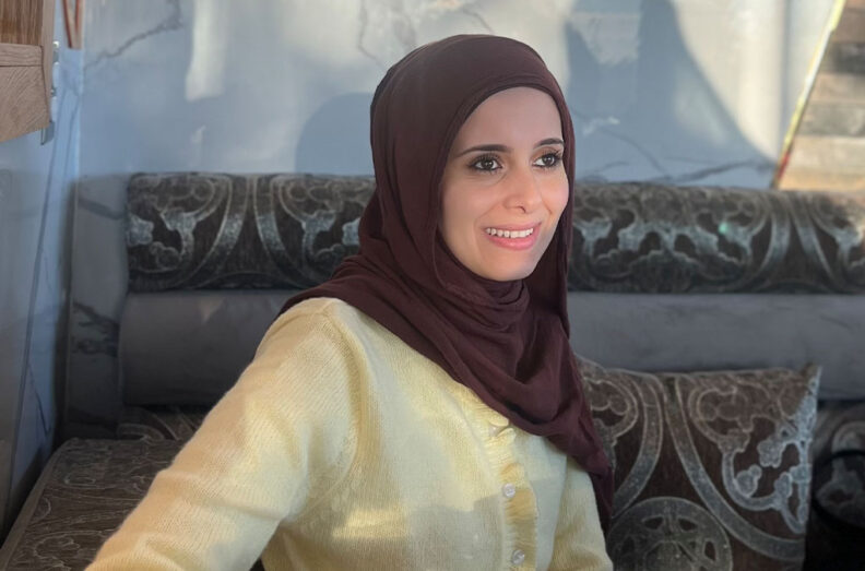 Sally AboHana wearing a maroon headscarf and light yellow buttoned sweater, seated on a patterned couch with a gray wall in the background.