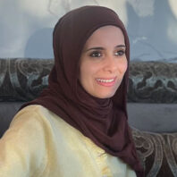 Sally AboHana wearing a maroon headscarf and light yellow buttoned sweater, seated on a patterned couch with a gray wall in the background.