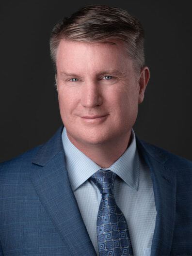 Shane Sullivan wearing a blue suit jacket and patterned tie against a dark background.