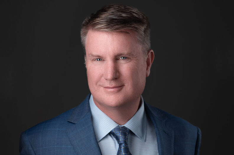 Shane Sullivan wearing a blue suit jacket and patterned tie against a dark background.