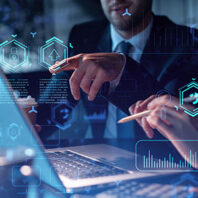 Two people collaborating at a desk with a laptop, pointing at the screen. Digital graphics of data charts, security icons, and upload symbols are overlaid, representing technology and analytics.