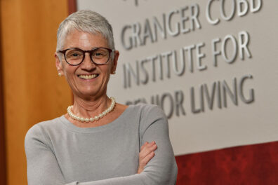 Nancy Swanger wearing a light gray top and pearl necklace, standing with arms crossed in front of a wall sign that reads “Granger Cobb Institute for Senior Living.”