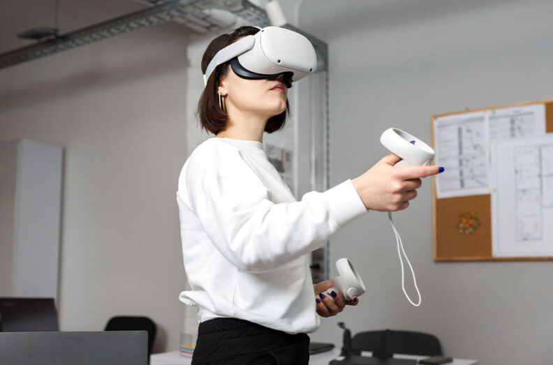 Person wearing a white sweatshirt using a virtual reality headset and handheld controllers in an office setting with a corkboard and papers in the background.