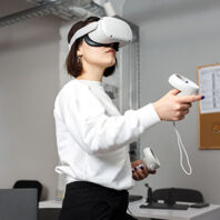 Person wearing a white sweatshirt using a virtual reality headset and handheld controllers in an office setting with a corkboard and papers in the background.