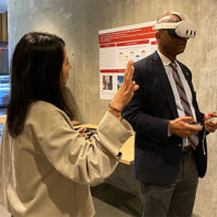 Two people interacting with virtual reality equipment in a modern room; one wearing a VR headset and holding controllers, while the other gestures with one hand. A red and white poster with diagrams is displayed on the concrete wall behind them.