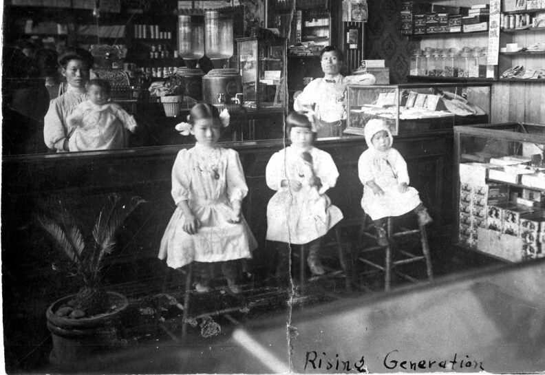 Historic photo of store counter with man and woman with baby and goods behind counter and three children in white dresses sitting on stools in front of counter