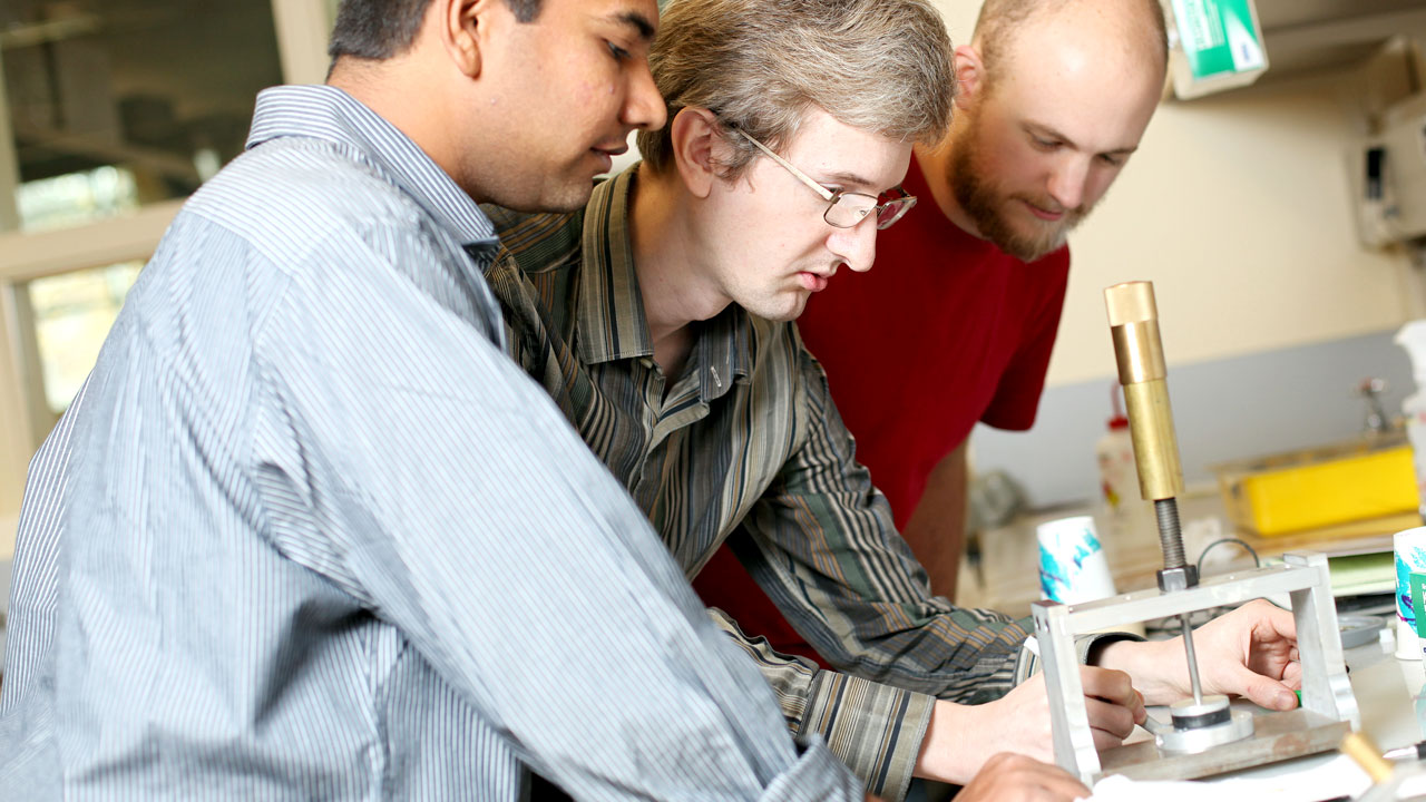 Two students observe a researcher manipulating a sample in a laboratory.