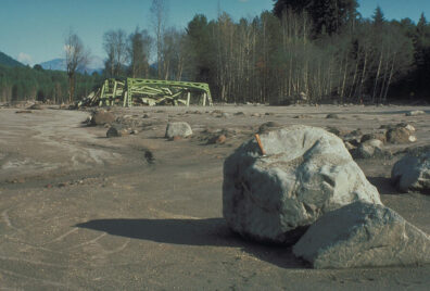 A damaged bridge is in the background. In the foreground are large rocks and debris.