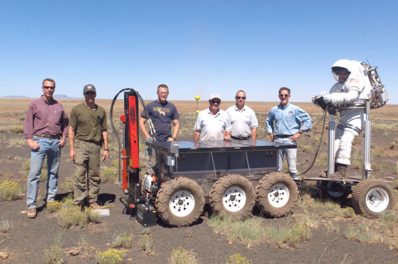 A group of men stand around a 6 wheeled lunar rover in the desert.