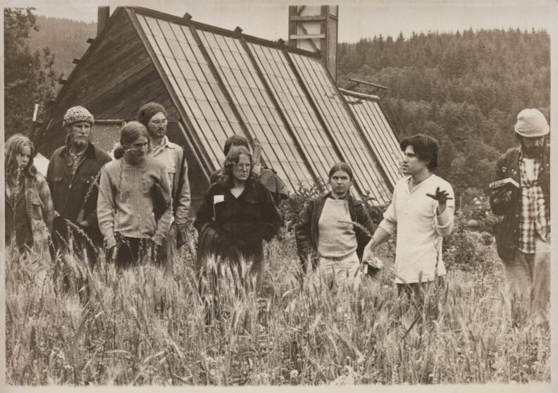 Black and white photo of several young people standing in a farm field with high grass. One person is speaking to them.