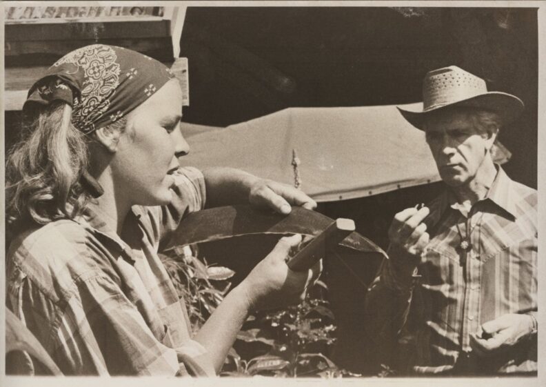 Black and white photo of a young woman wearing a bandana on her head and holding a scythe while an older man wearing a cowboy hat stands nearby looking at the scythe.