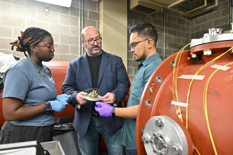 Three individuals standing in a lab with large metal equipment in conversation.