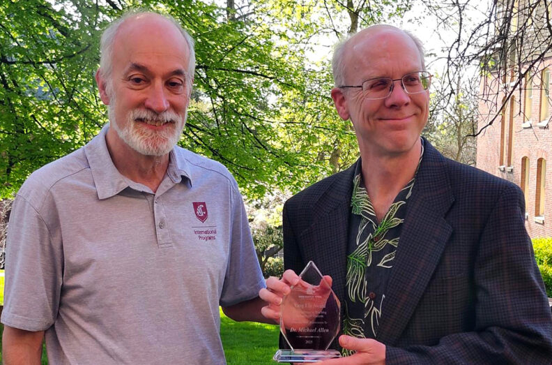 Two men stand side by side with smiles, as one holds an award.