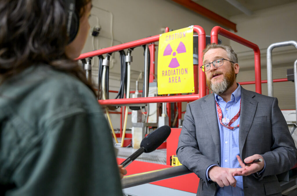 Corey Hines speaks to a journalist wearing headphones and holding a microphone during a tour of WSU's Nuclear Science Center.