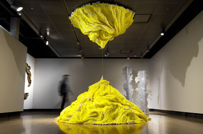 A large, bright yellow abstract sculpture with a suspended, draped form hanging from the ceiling and connecting to a matching mound on the floor.
