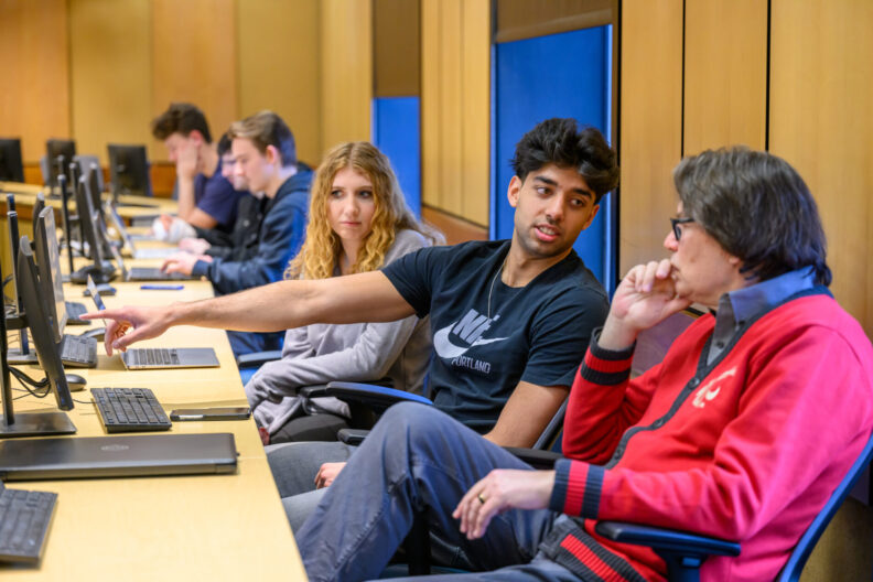 Students and instructor discuss work in a computer lab. The student in the foreground is pointing to a computer screen while explaining.