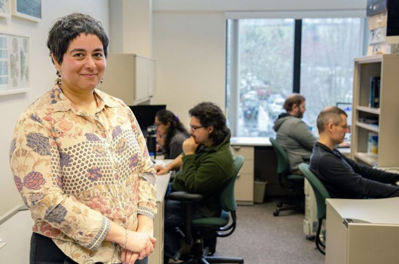 A closeup of Deepti Singh standing in a research lab as four students sit and work at computer stations behind her.