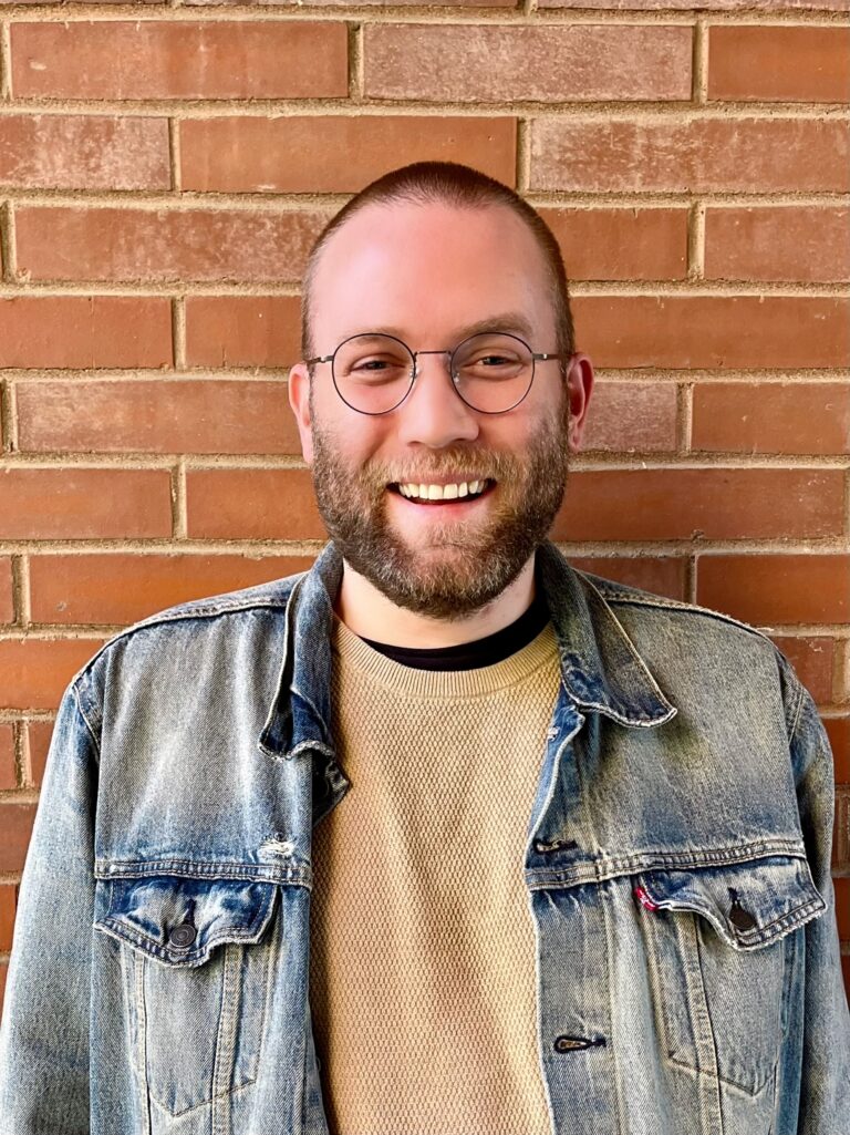 A man wearing glasses and a denim jacket stands in front of a brick wall.
