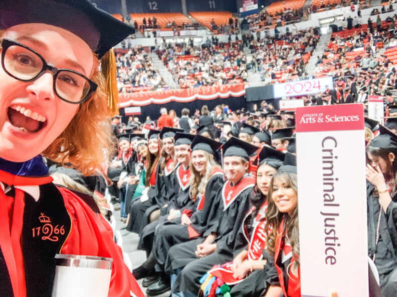 Selfie of Melanie-Angela Neuilly in stadium filled with undergraduates in caps and gowns and Criminal Justice sign. 