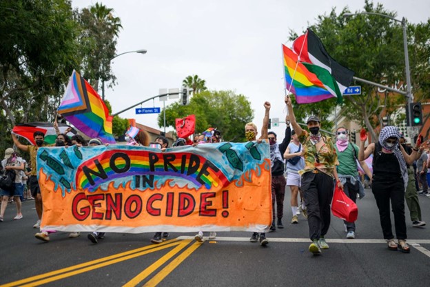 A protest march that includes people carrying LGBTQ+ flag. In the foreground, participants in the march carry a large banner with the phrase, "No Pride in Genocide".