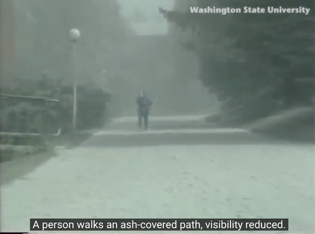 Video still showing a person walking on a pedestrian pathway covered in ash. "Washington State University" is printed in the top right, and a caption below reads, "A person walks an ash-covered path, visibility reduced."