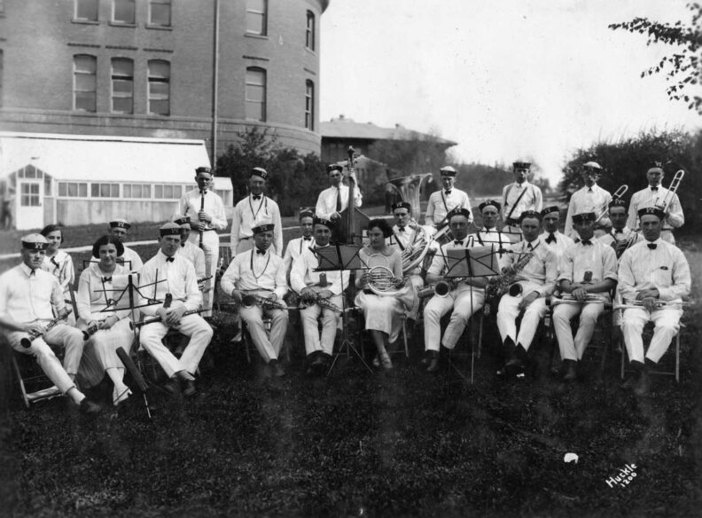 One b&w photographic print of a WSC band wearing uniforms and sitting with instruments for Campus Day, with the curved edge of Science (later Murrow) Hall behind them.
