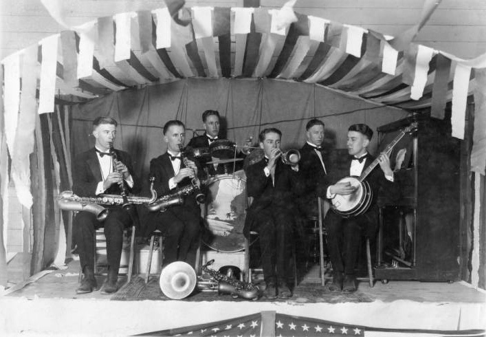 Small jazz band on stage with two saxophonists doubling up on clarinets, a trumpet player with French horn at his feet, a banjo, piano, and drum set.