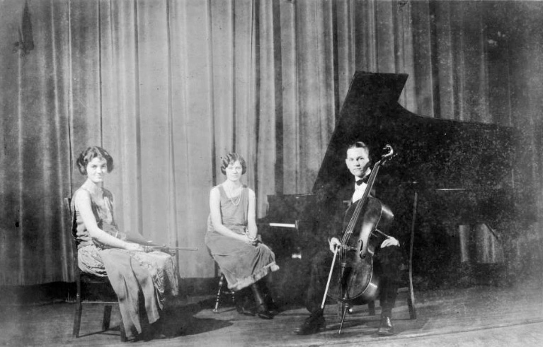 Musical trio sitting on stage with instruments in resting position. One with violin, one at the piano, and one with cello.