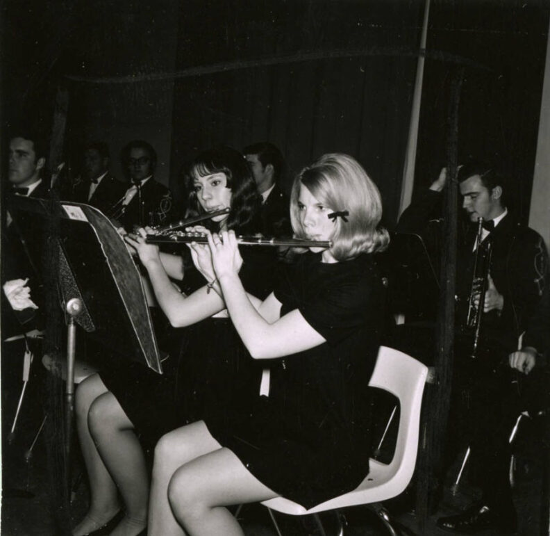 Symphonic band concert with musicians, wearing concert black, sitting in chairs on stage. Flutists in front with trumpet player and others in background.