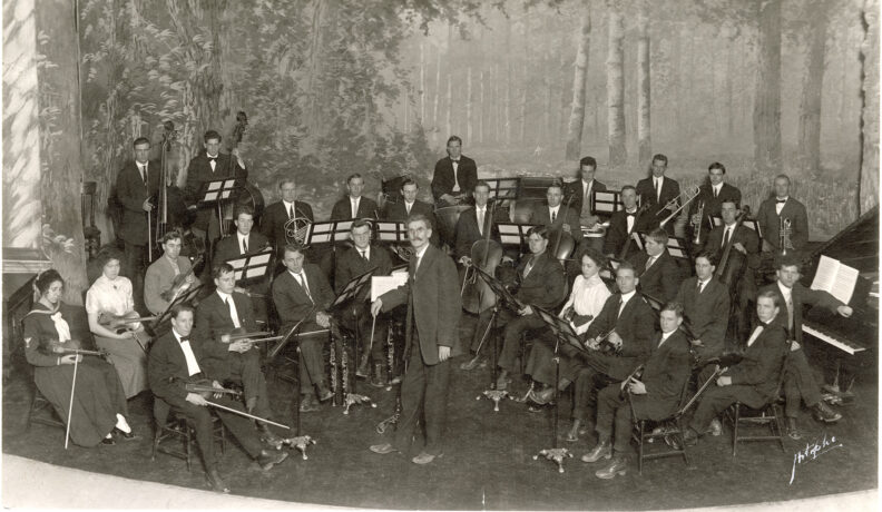 Orchestral players and conductor on stage with painted backdrop of outdoor forest scene