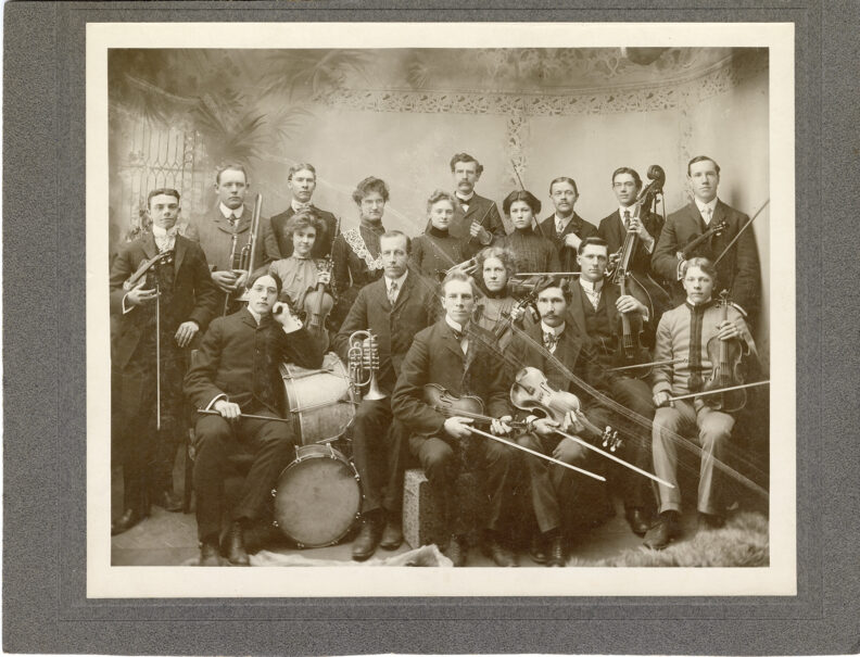Historical photo of predominantly string players, a percussionist and a few brass players, holding instruments.